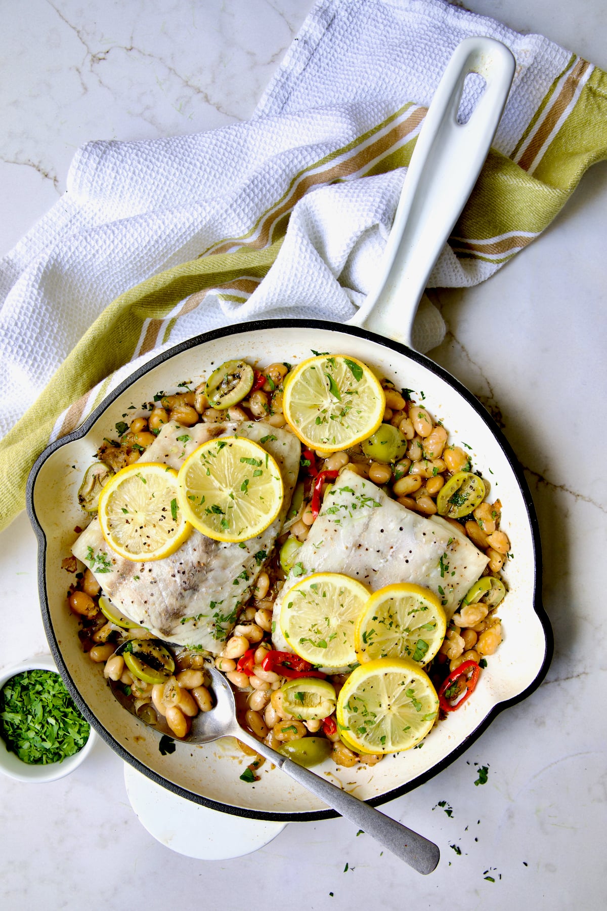 Mediterranean Fish and White Bean Skillet in white cast iron skillet topped with lemon slices sprinkled with fresh parsley.