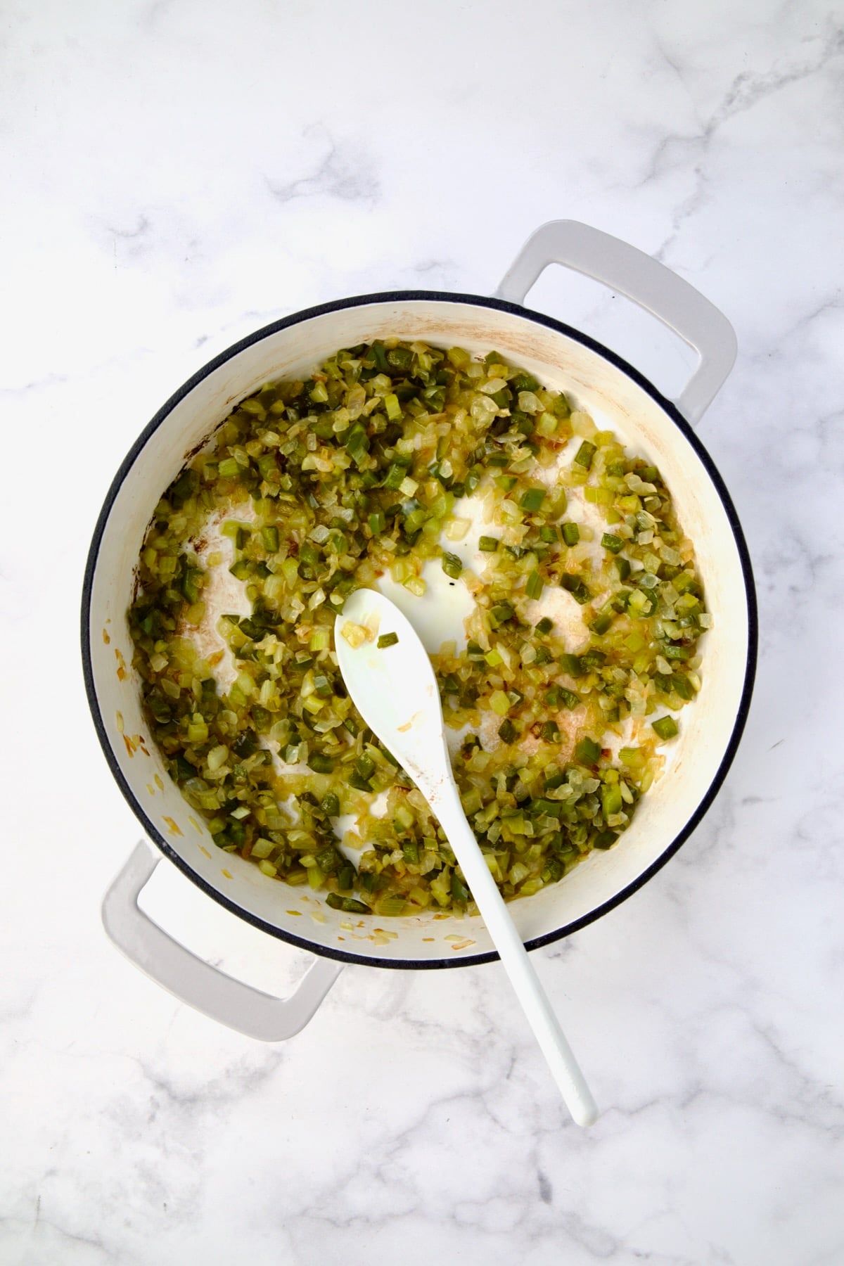Cooked onions, celery and green bell pepper being sauteed in white enamel cast iron skillet with mixing spoon.
