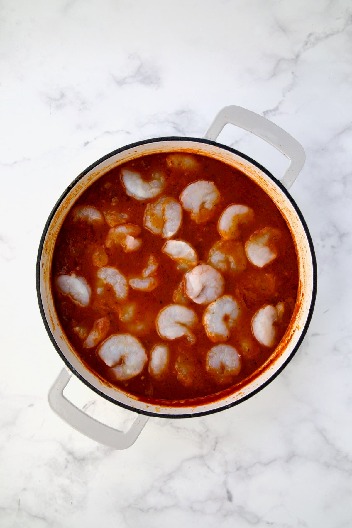 Uncooked shrimp added to tomato sauce in white enamel cast iron skillet.