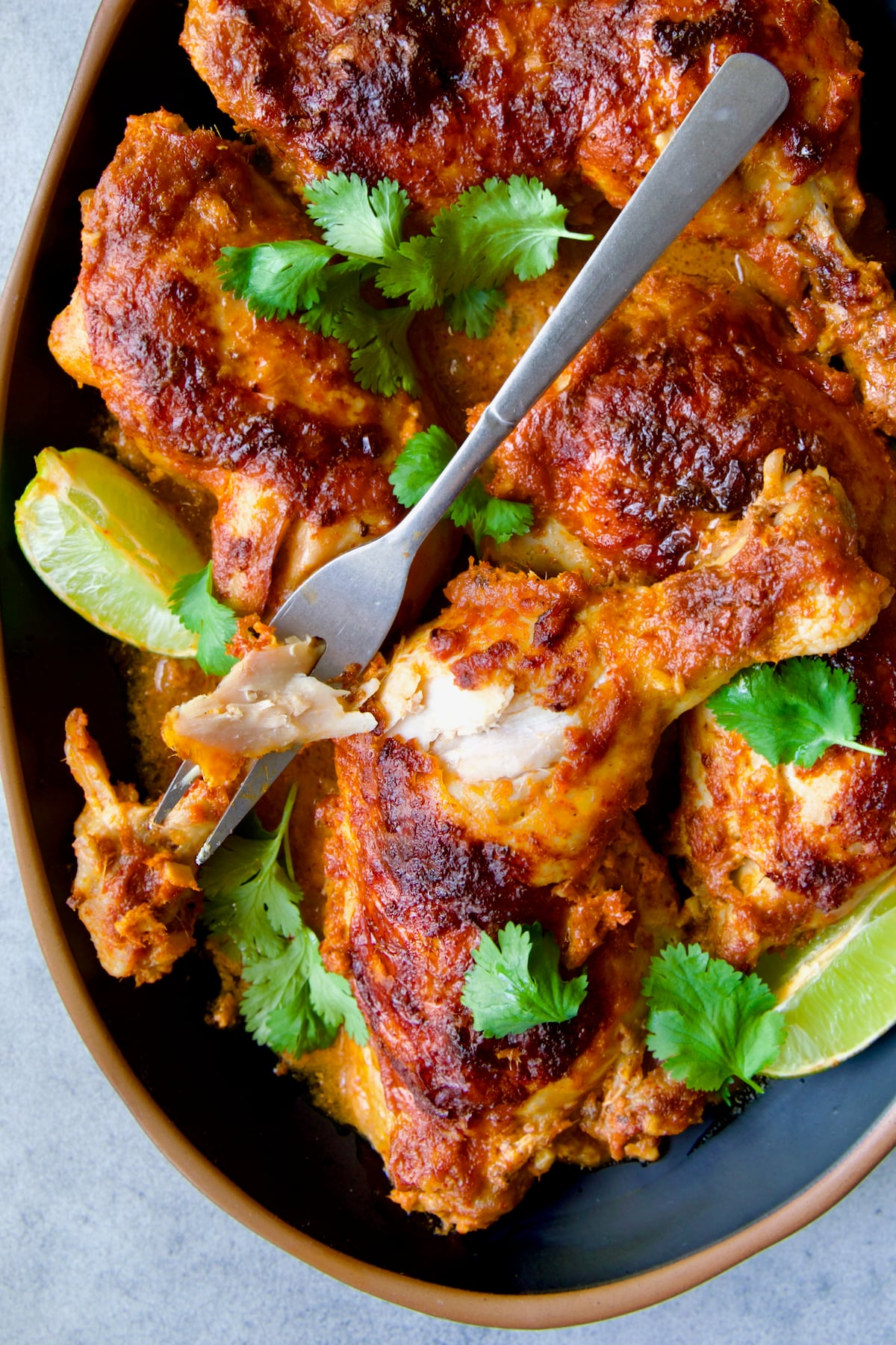 Thai Red Curry Braised Chicken Legs on black platter garnished with cilantro and lime wedges with serving fork.