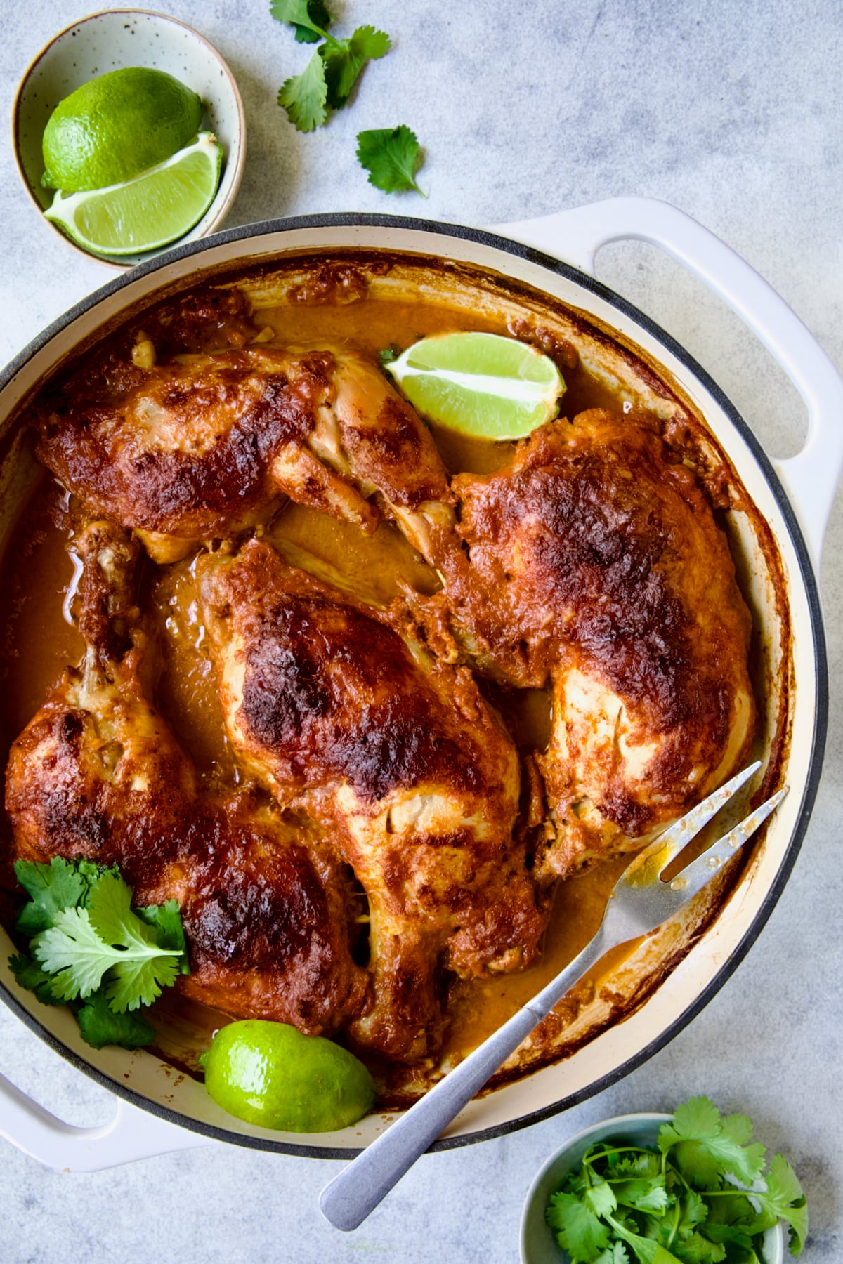 Thai Red Curry Braised Chicken Legs in white enamel cast iron skillet garnished with cilantro and lime wedges with serving fork.