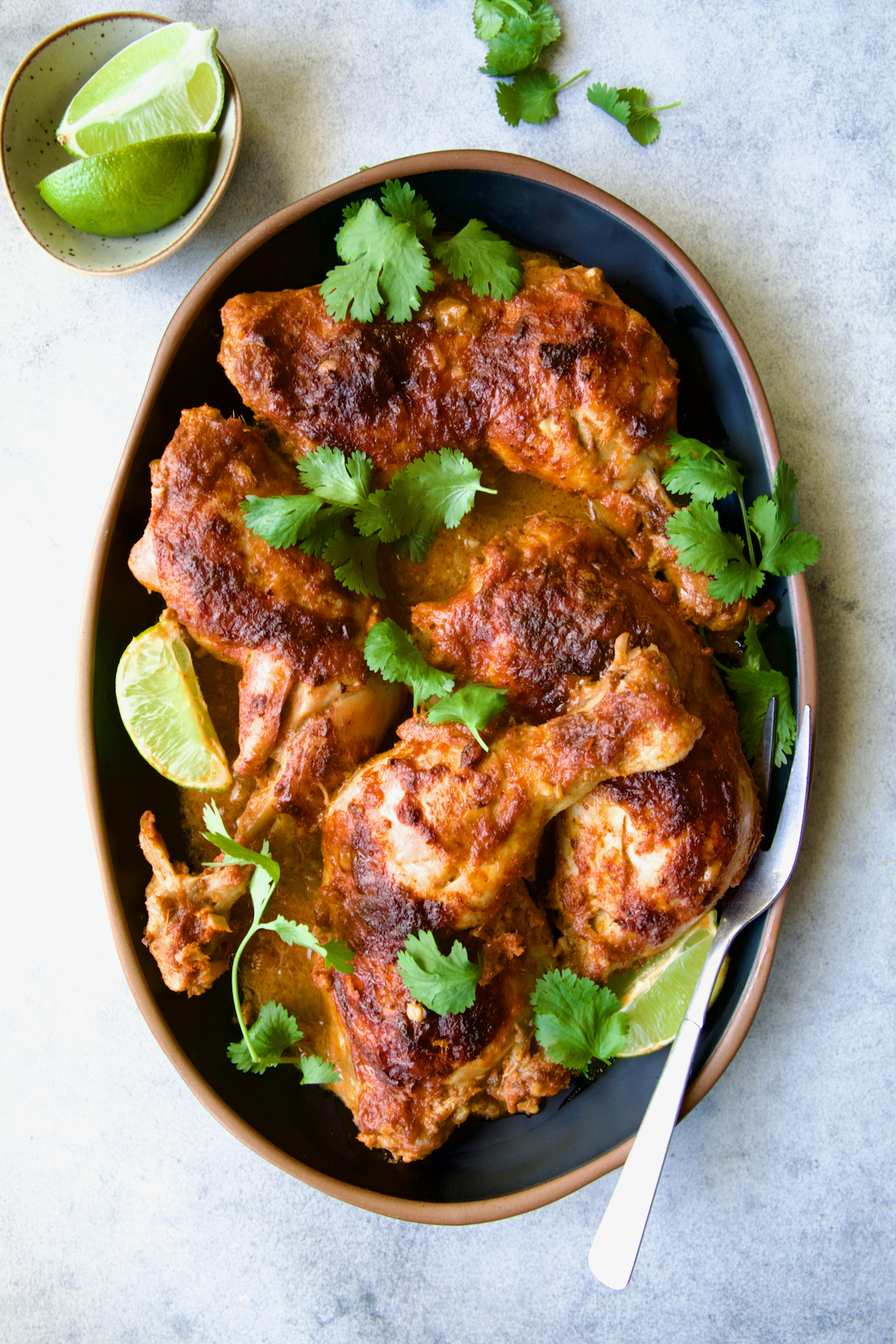Thai Red Curry Braised Chicken Legs on black platter garnished with cilantro and lime wedges with serving fork.