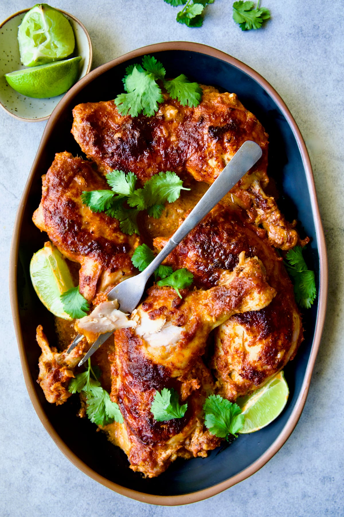 Thai Red Curry Braised Chicken Legs on black platter garnished with cilantro and lime wedges with serving fork.