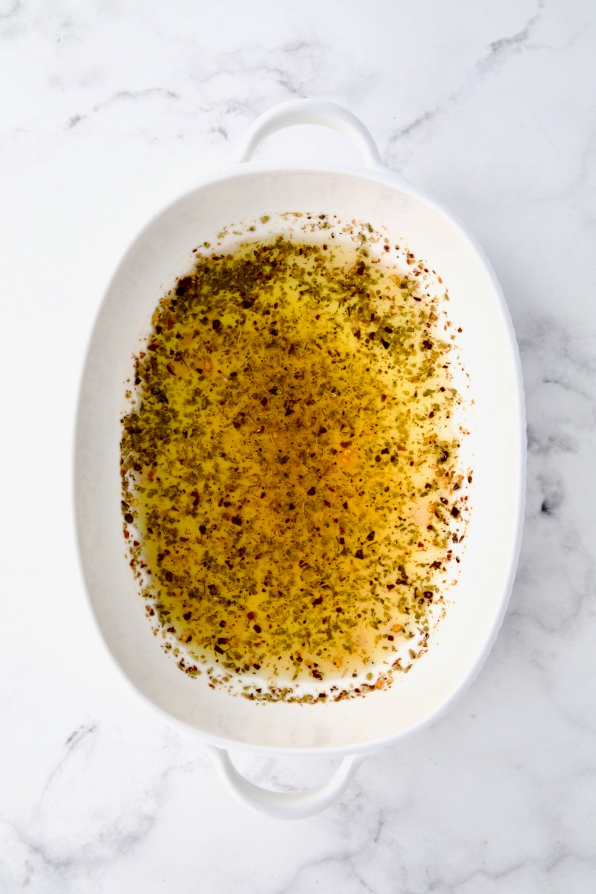 Olive oil, lemon juice, dried oregano and crushed red pepper flakes mixed together in white oval baking dish.