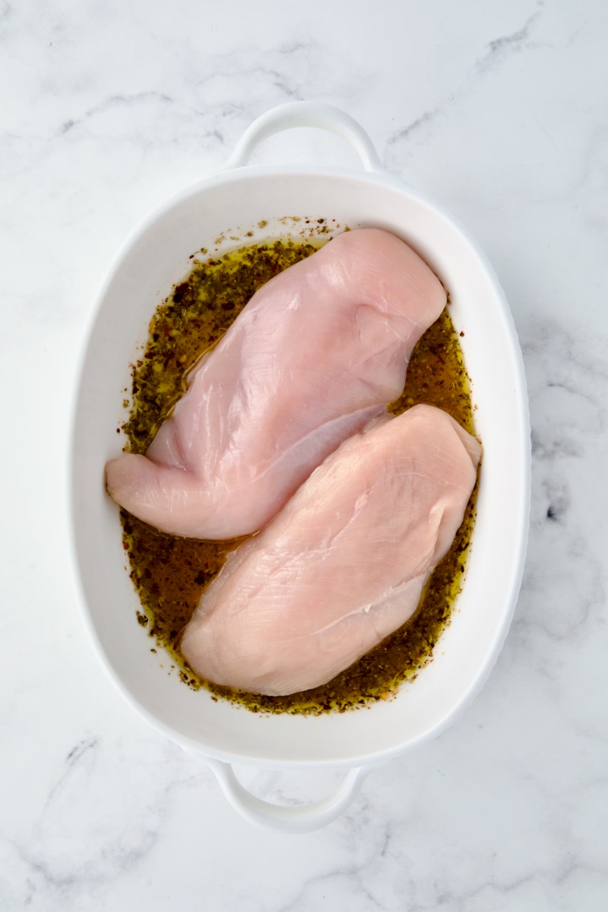 Two uncooked chicken breasts in white oval baking dish atop herb, lemon and olive oil combination.