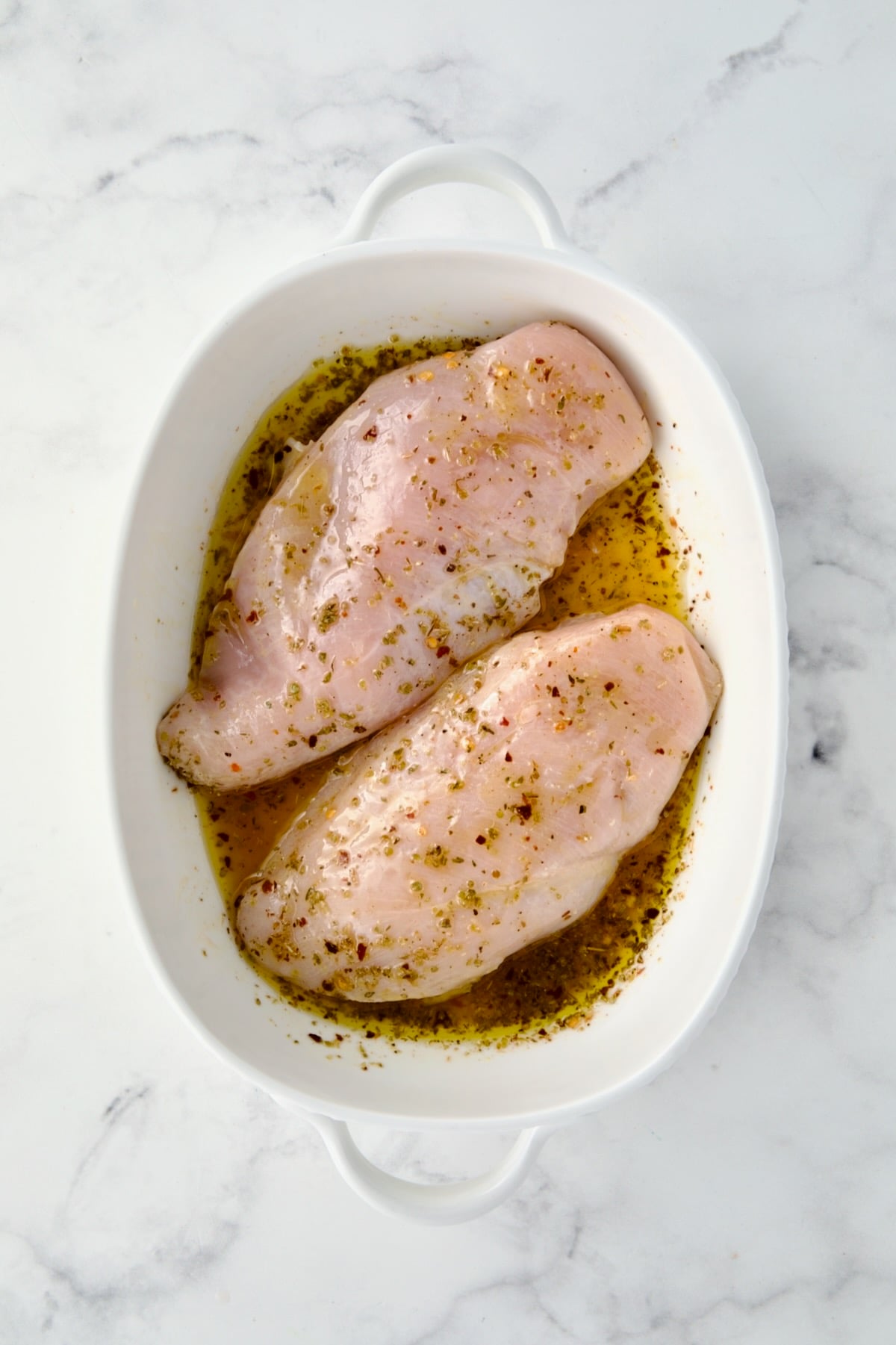 Two uncooked chicken breasts in white oval baking dish topped with herb, lemon and olive oil combination.