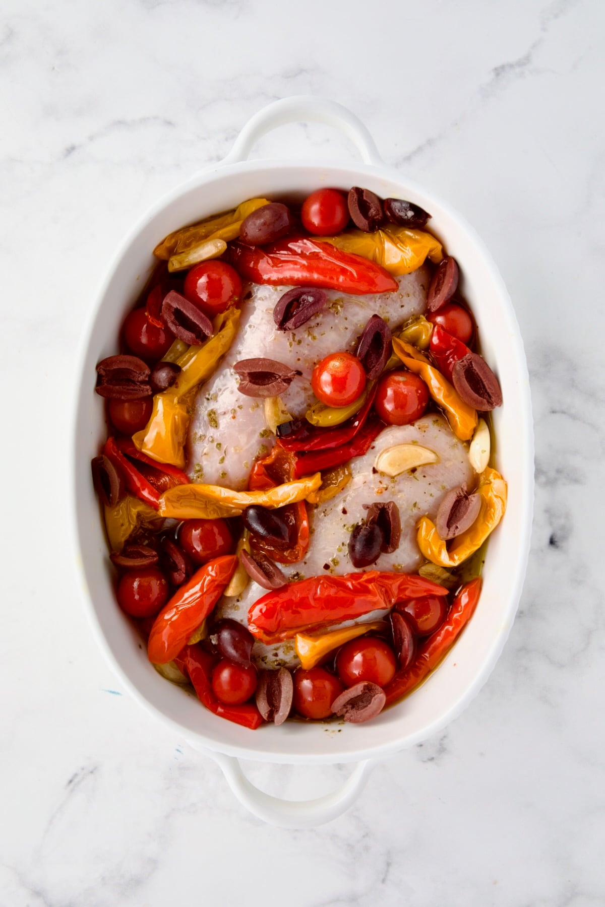 Two uncooked chicken breasts in white oval baking dish with roasted red peppers, olives, tomatoes and garlic added.