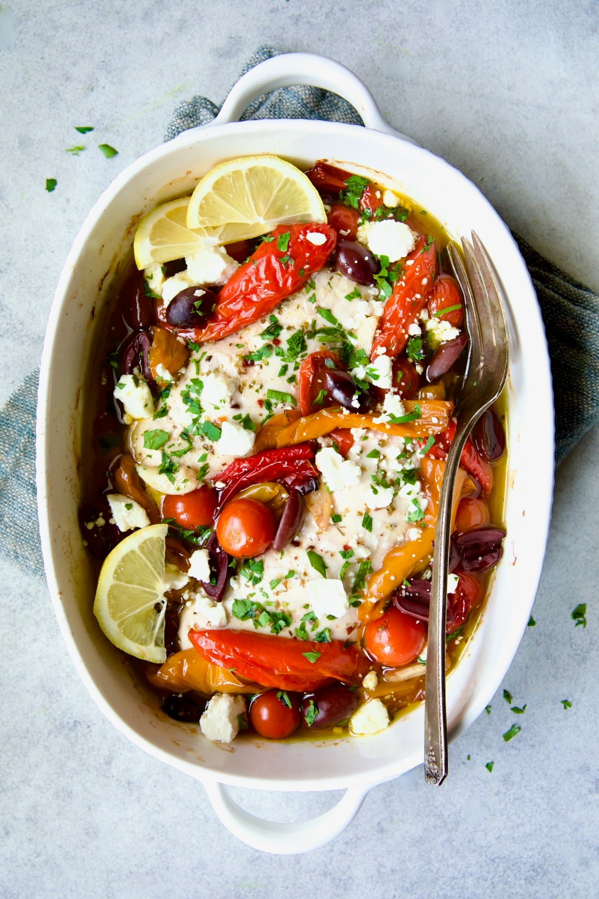 Greek Baked Chicken in white oval baking dish with serving fork on blue napkin.