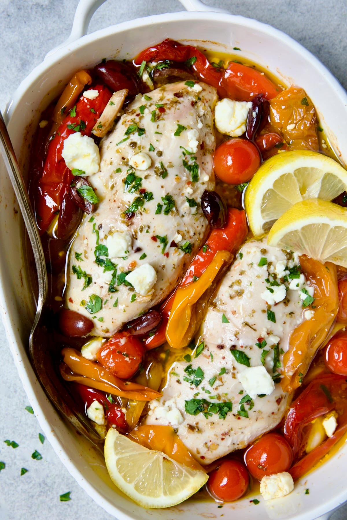 Greek Baked Chicken in white oval baking dish with serving fork garnished with lemon wedges.