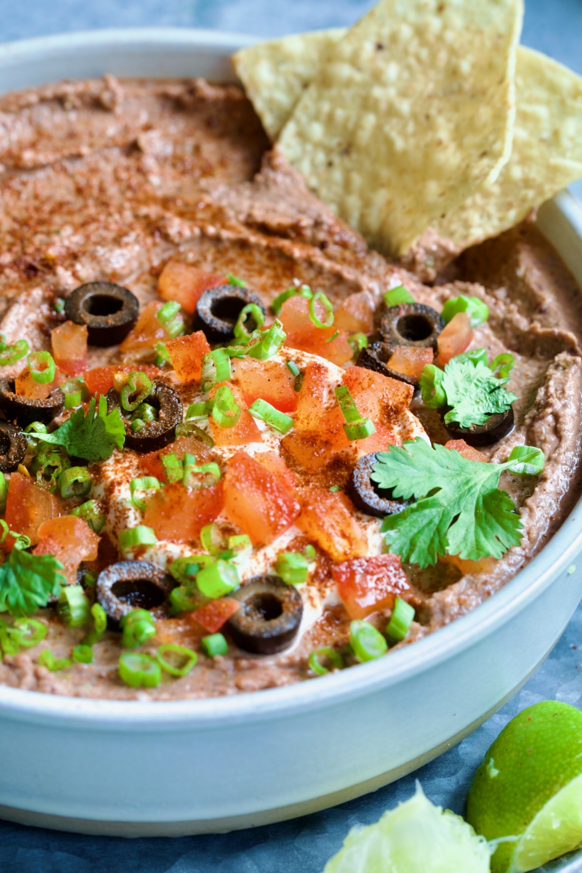 Black Bean Dip garnished with black olives, tomatoes, scallions and cilantro leaves in white clay bowl with tortilla chips and lime wedges.