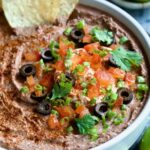 Black Bean Dip garnished with black olives, tomatoes, scallions and cilantro leaves in white clay bowl with tortilla chips and lime wedges.