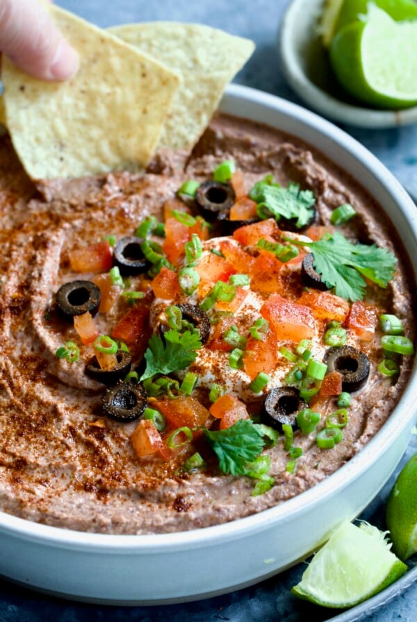 Black Bean Dip garnished with black olives, tomatoes, scallions and cilantro leaves in white clay bowl with tortilla chips and lime wedges.