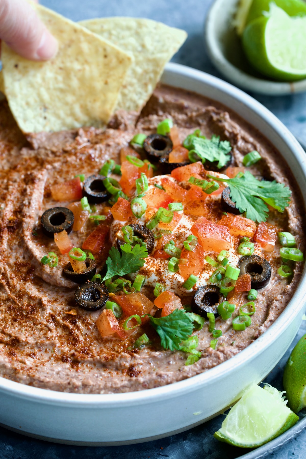 Black Bean Dip garnished with black olives, tomatoes, scallions and cilantro leaves in white clay bowl with tortilla chips and lime wedges.