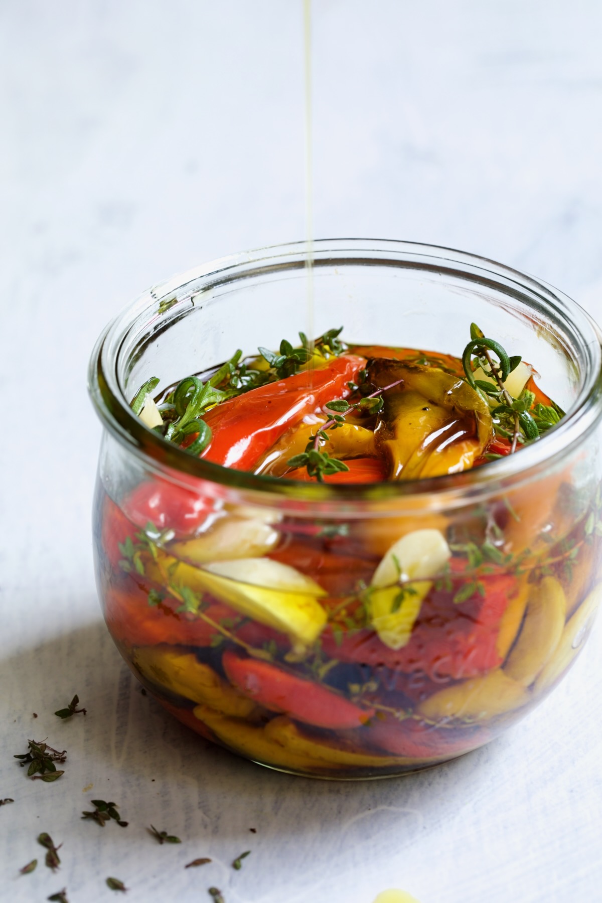 Roasted Mini Peppers in glass jar with fresh herbs and garlic with olive oil being drizzled in.