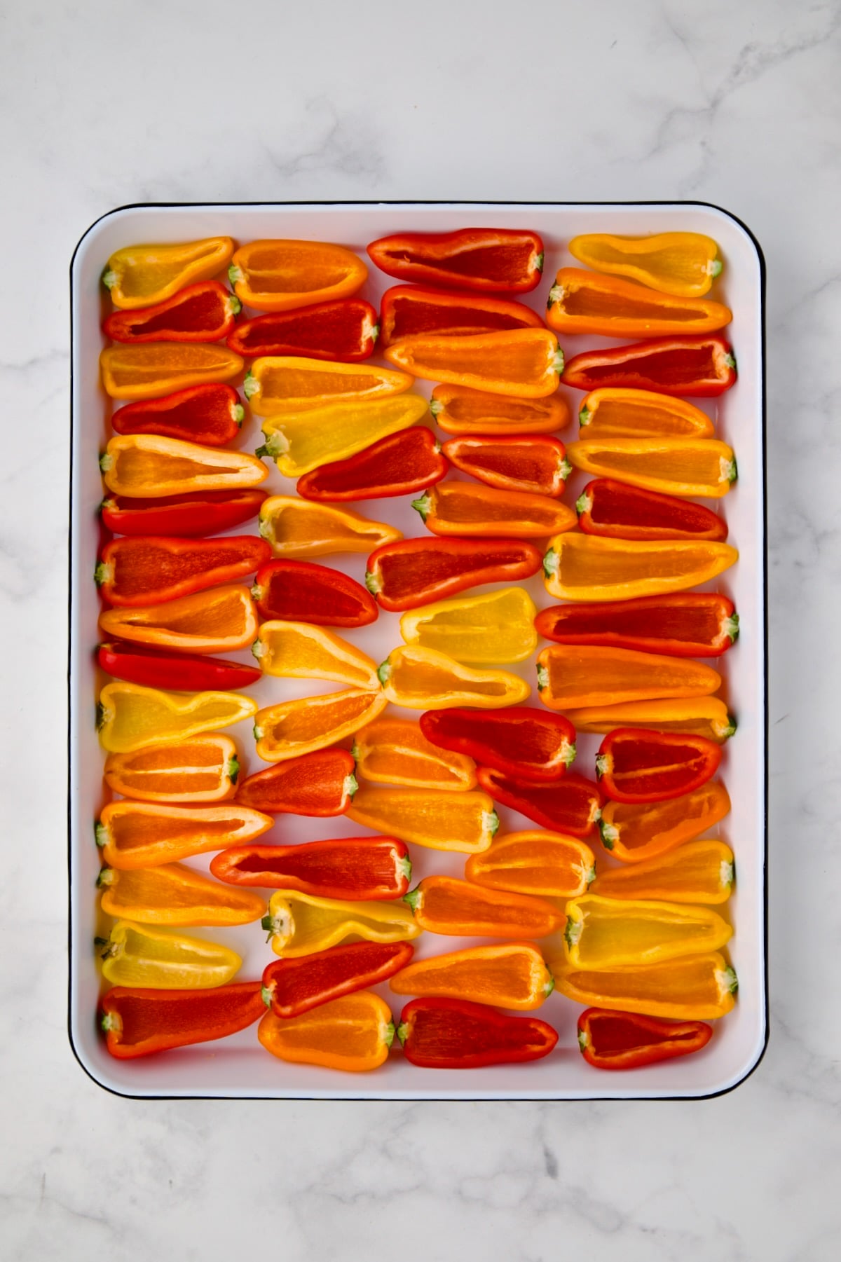 Halved mini peppers on white enamel rimmed baking sheet.