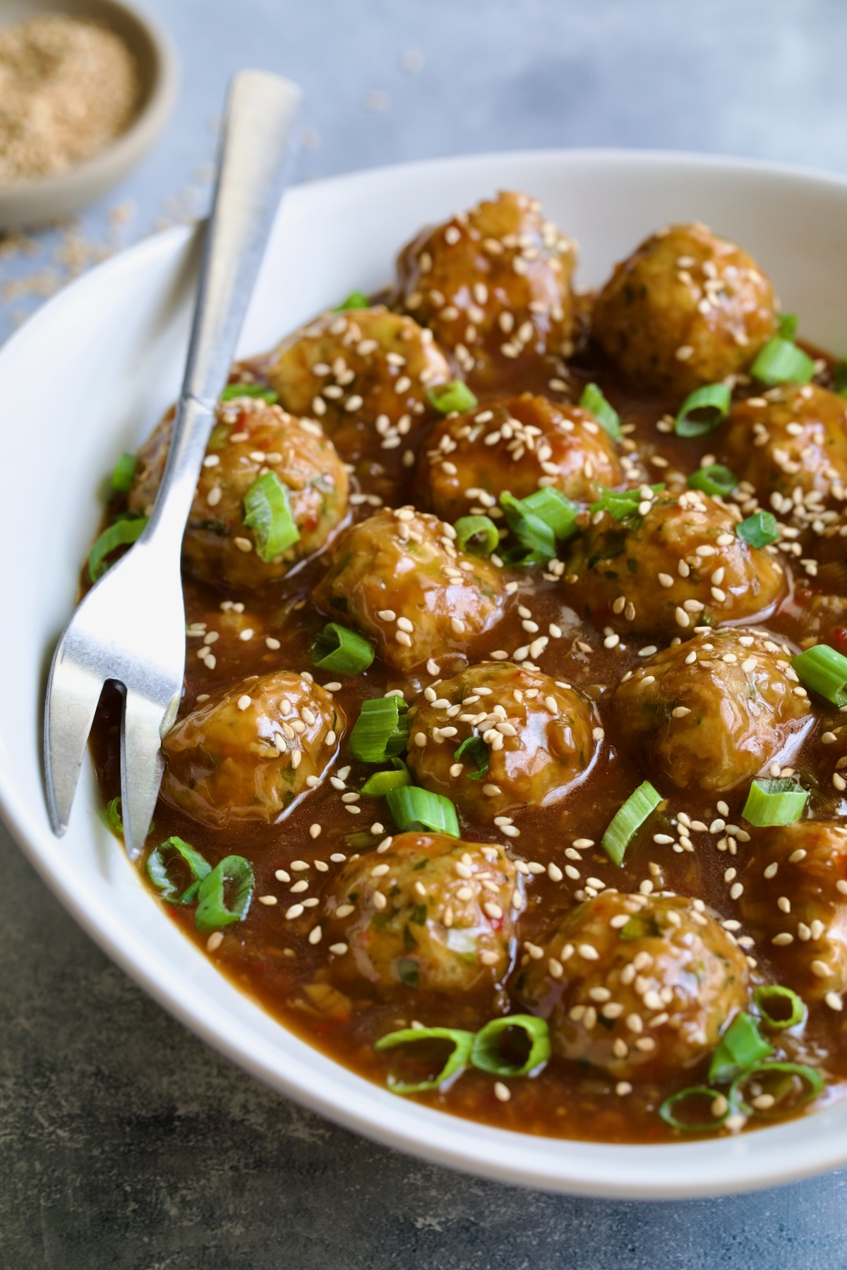 Asian Turkey Meatballs in white oval serving bowl with serving fork garnished with sesame seeds and scallions.