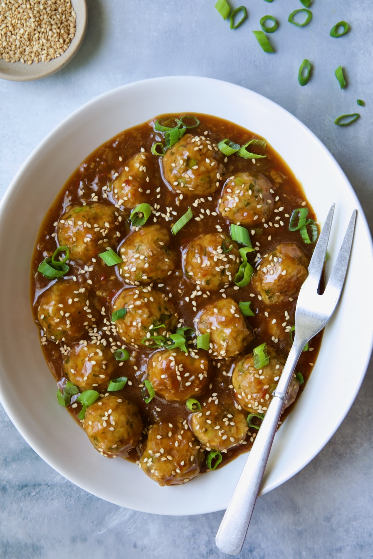 Asian Turkey Meatballs in white oval serving bowl with serving fork garnished with sesame seeds and scallions.