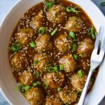 Asian Turkey Meatballs in white oval serving bowl with serving fork garnished with sesame seeds and scallions.