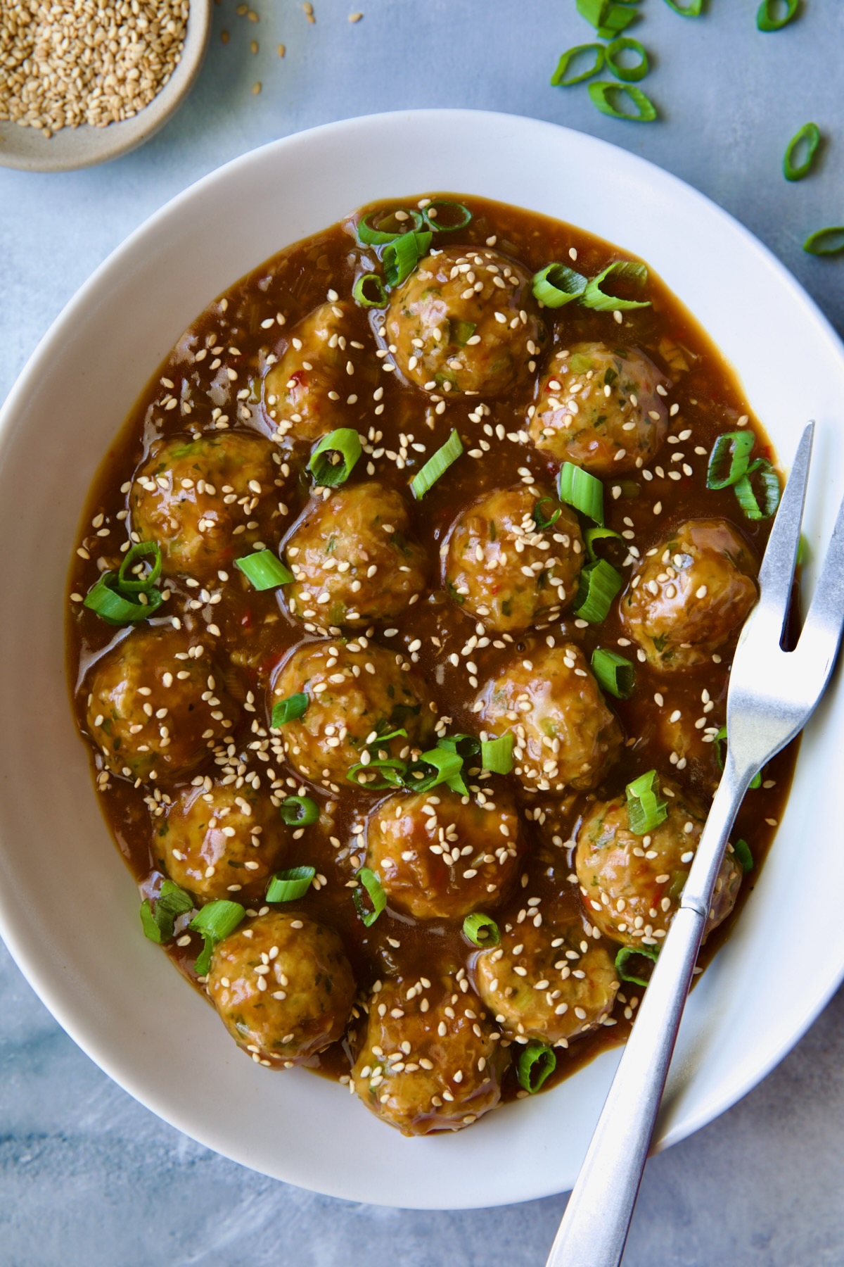 Asian Turkey Meatballs in white oval serving bowl with serving fork garnished with sesame seeds and scallions.