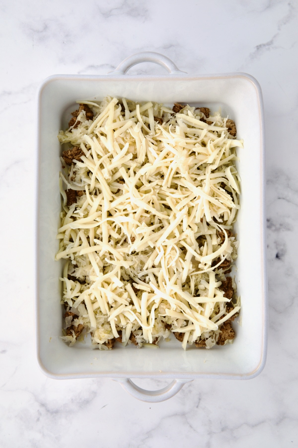 Cheese placed over rye bread cubes and sauerkraut in white rectangular baking dish.