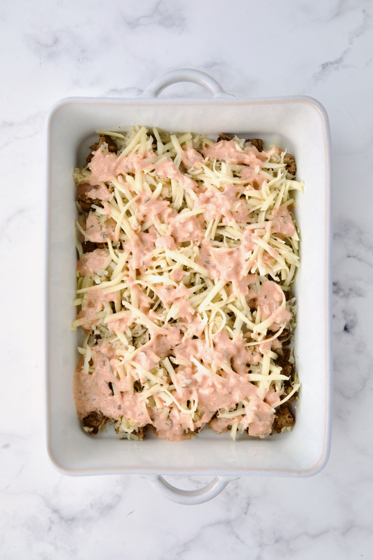 Thousand Island dressing drizzled over rye bread cubes, sauerkraut and Swiss cheese in white rectangular baking dish.