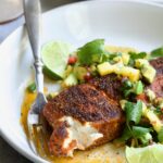 Blackened Halibut with Mango - Avocado Salsa on white plate with fork garnished with cilantro and lime wedge with a piece removed to show the inside of the fish.