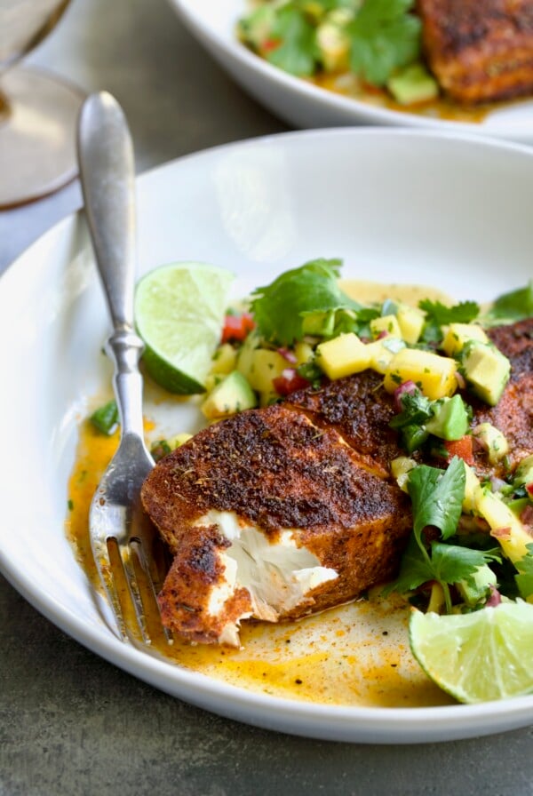 Blackened Halibut with Mango - Avocado Salsa on white plate with fork garnished with cilantro and lime wedge with a piece removed to show the inside of the fish.