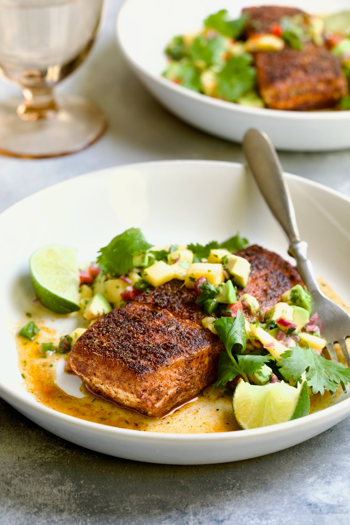 One serving of Blackened Halibut with Mango - Avocado Salsa on white plate with fork garnished with cilantro and lime wedges.