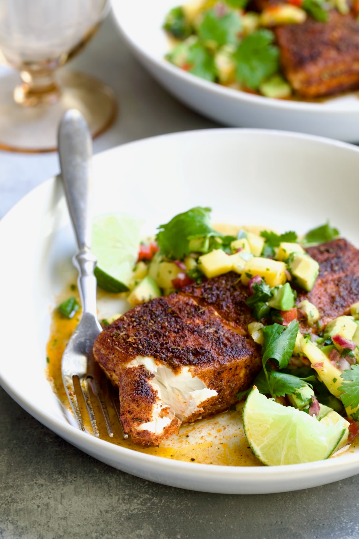 One serving of Blackened Halibut with Mango - Avocado Salsa on white plate with fork garnished with cilantro and lime wedges with piece flaked away to show the interior.