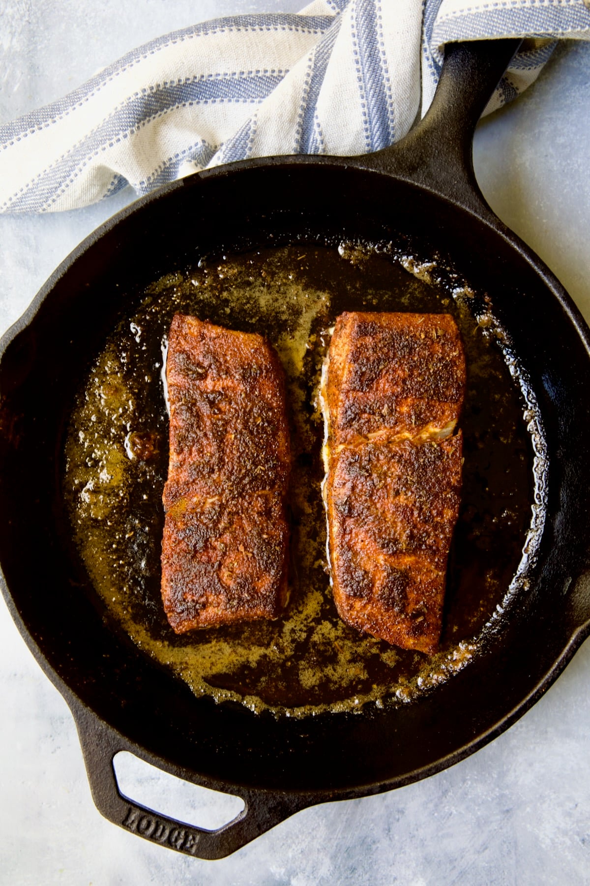 Two pieces of halibut coated with blackening seasoning in cast iron skillet after being cooked with blue striped towel wrapped around the handle.