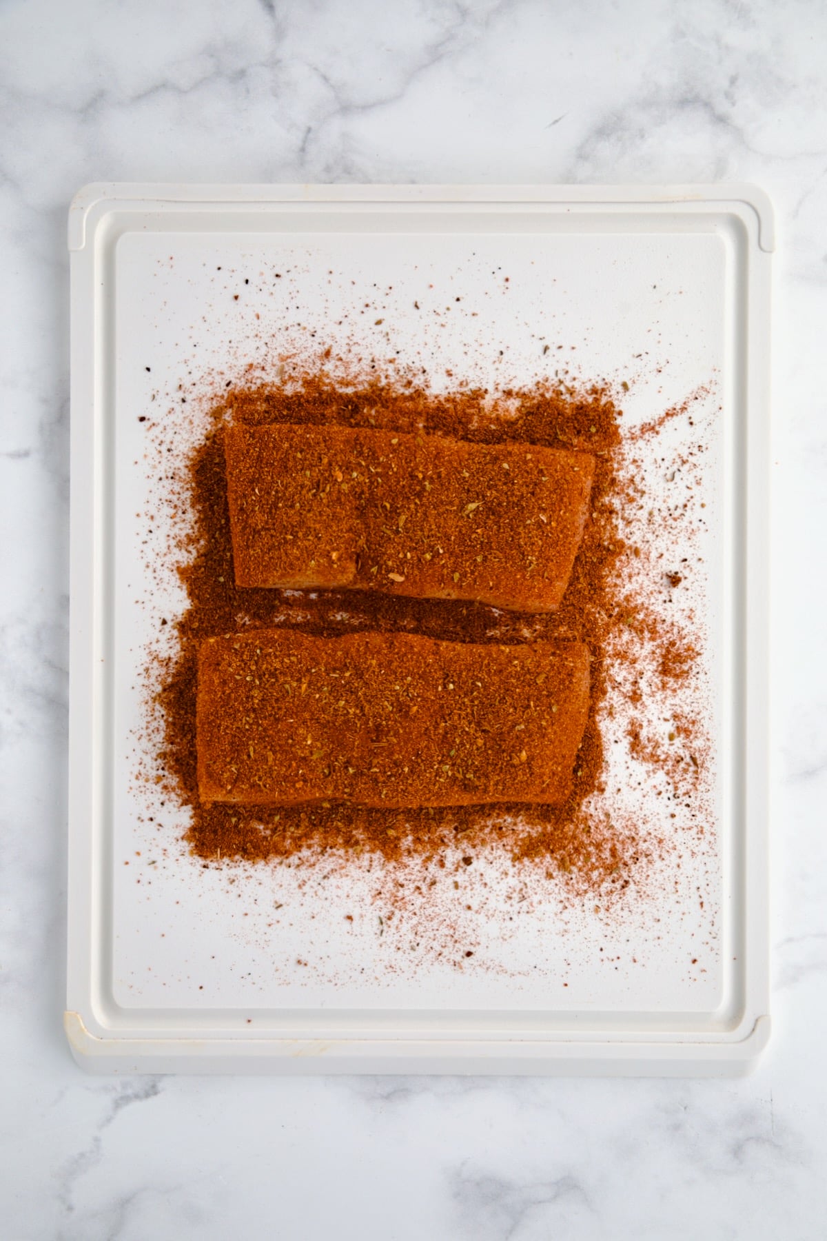 Two pieces of uncooked halibut coated with blackening seasoning on white plastic cutting board.