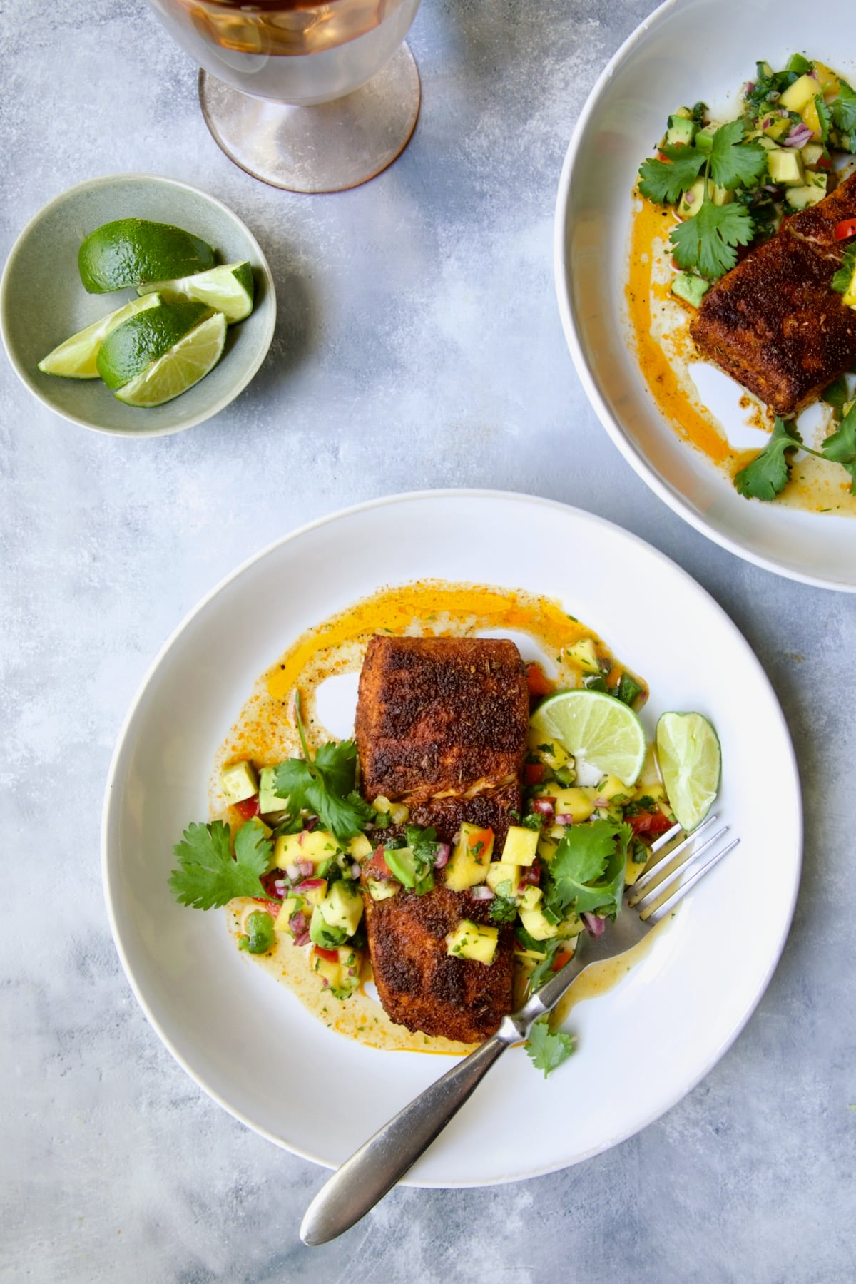 Two servings of Blackened Halibut with Mango - Avocado Salsa on white plates garnished with cilantro and lime wedge.