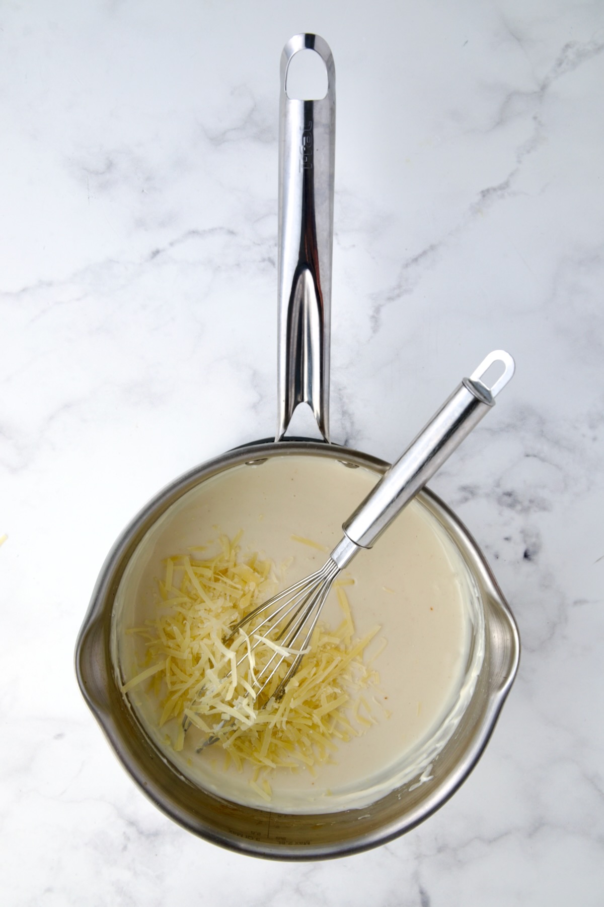 Cheese being added to cream sauce in stainless steel saucepan.