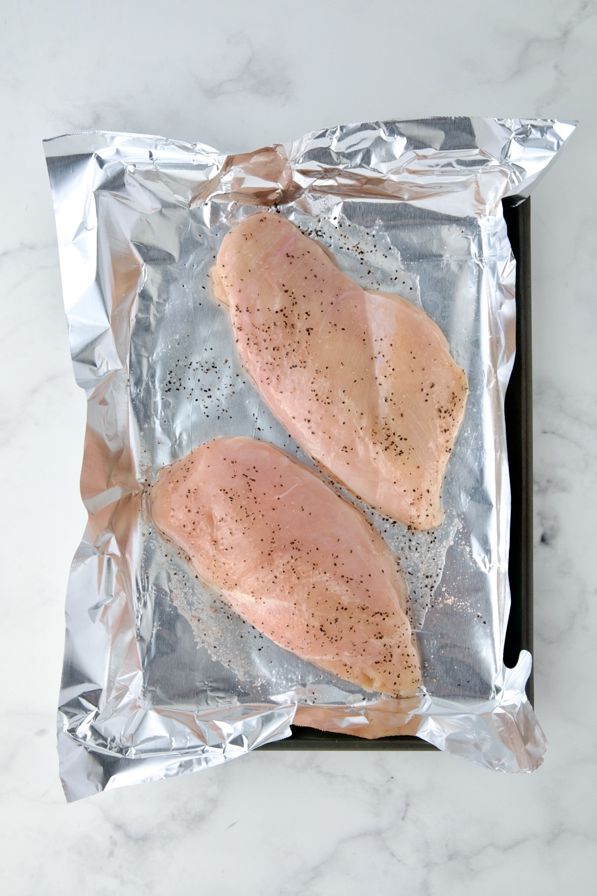 Two uncooked chicken breasts on aluminum foil lined baking sheet sprinkled with salt and black pepper.