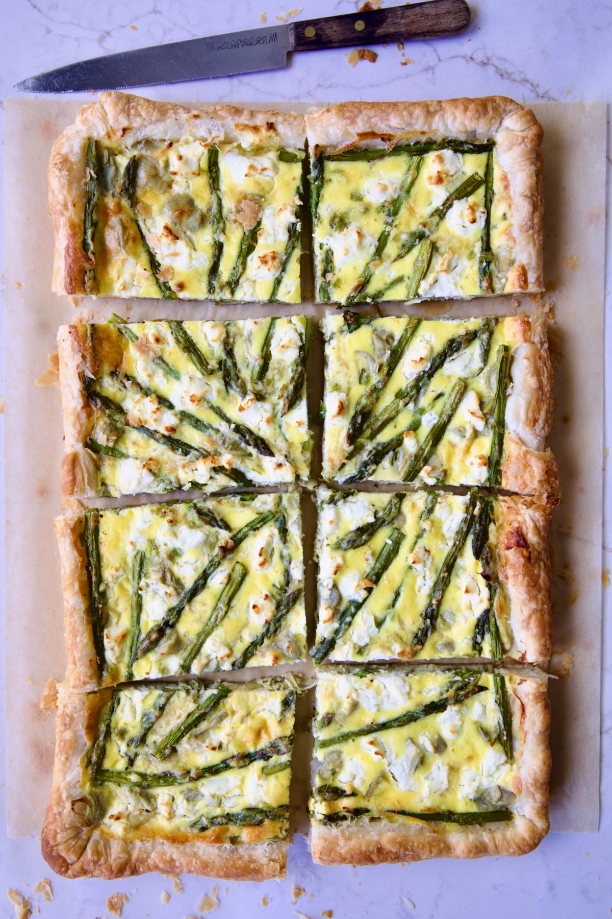 Puff Pastry Vegetable Tart on brown parchment paper cut into eight pieces with knife nearby.