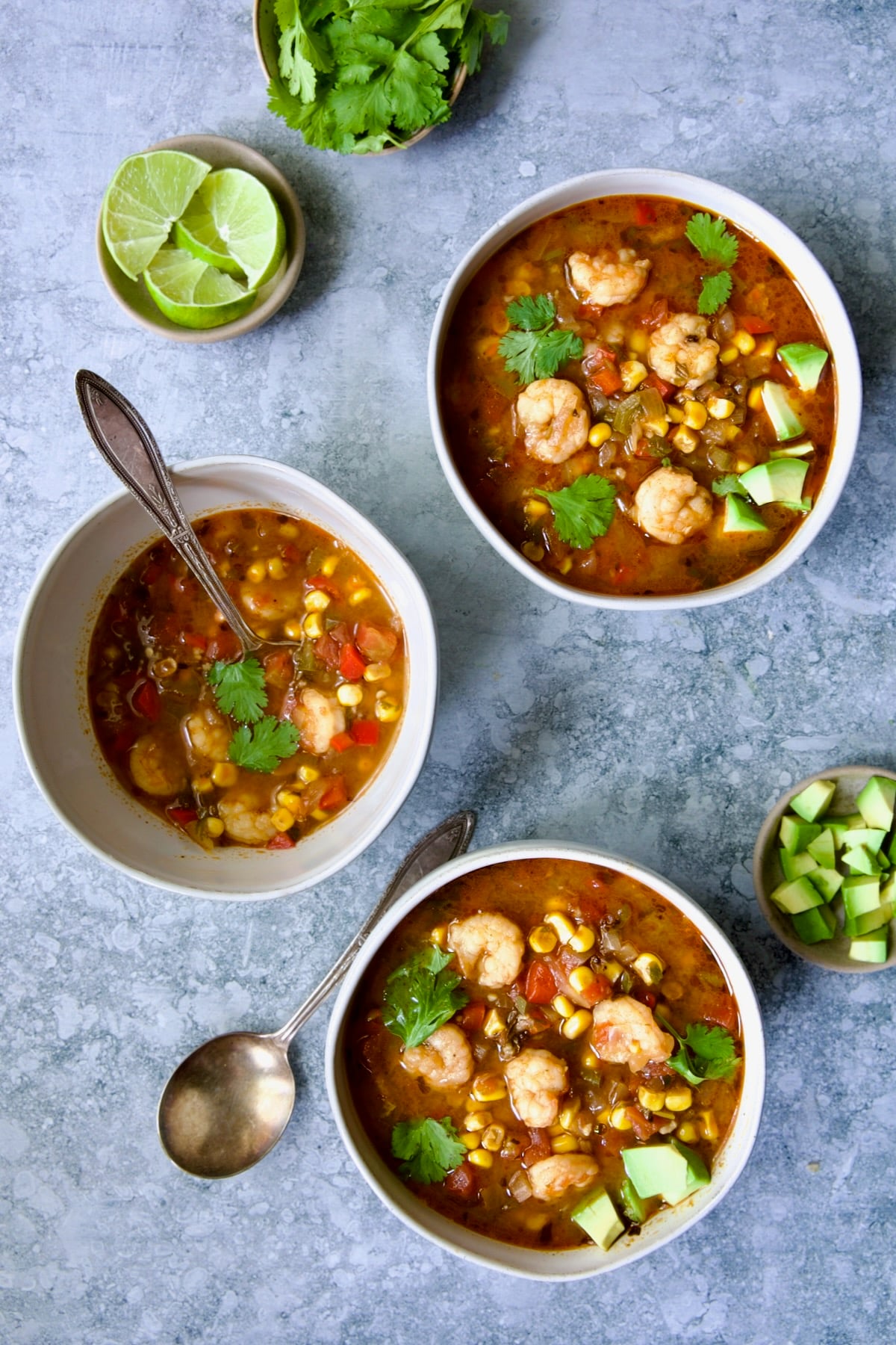Three bowls of Shrimp and Corn Soup with smaller bowls of lime wedges, cilantro and diced avocado.