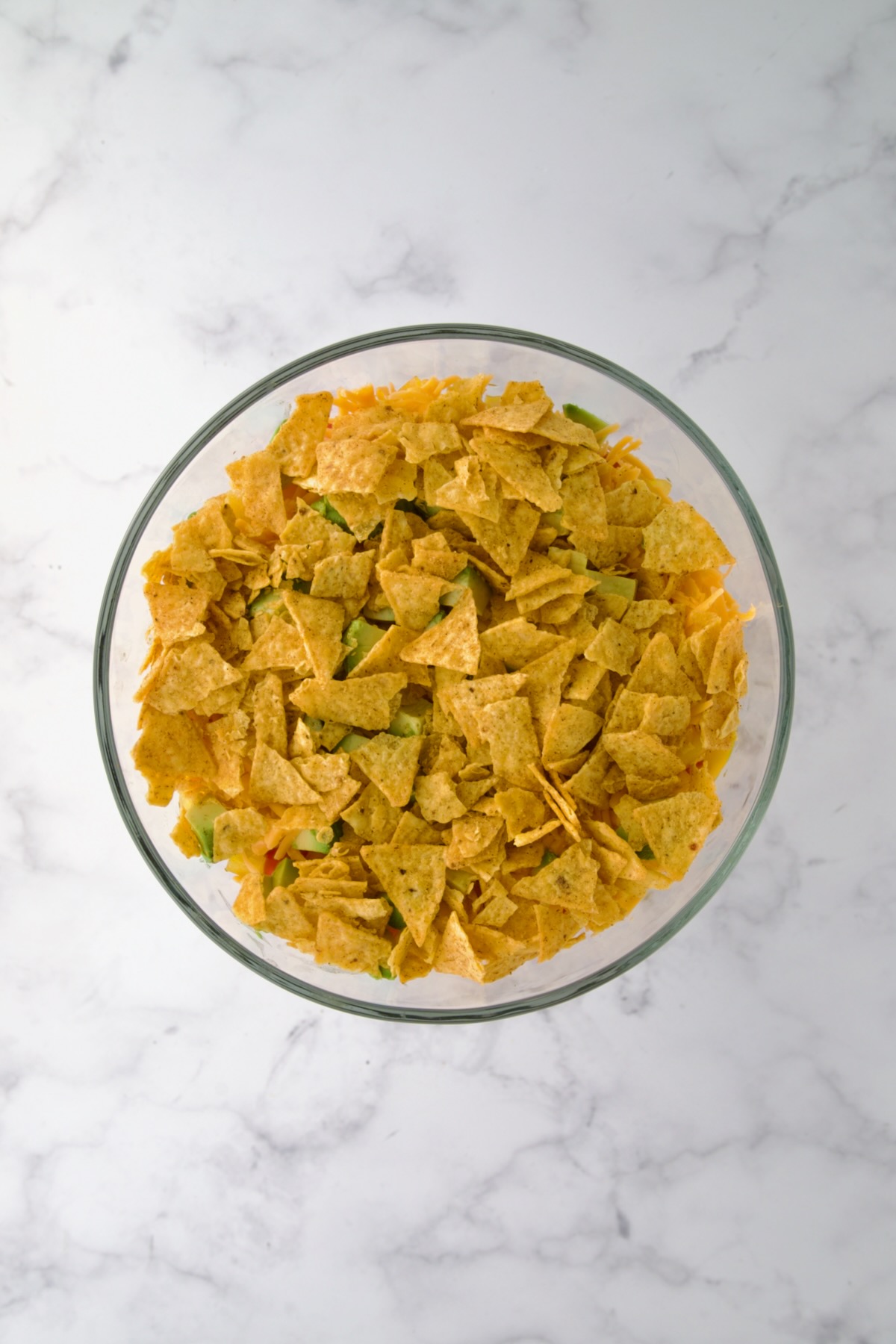 Crushed tortilla chips over avocado in glass footed serving bowl.