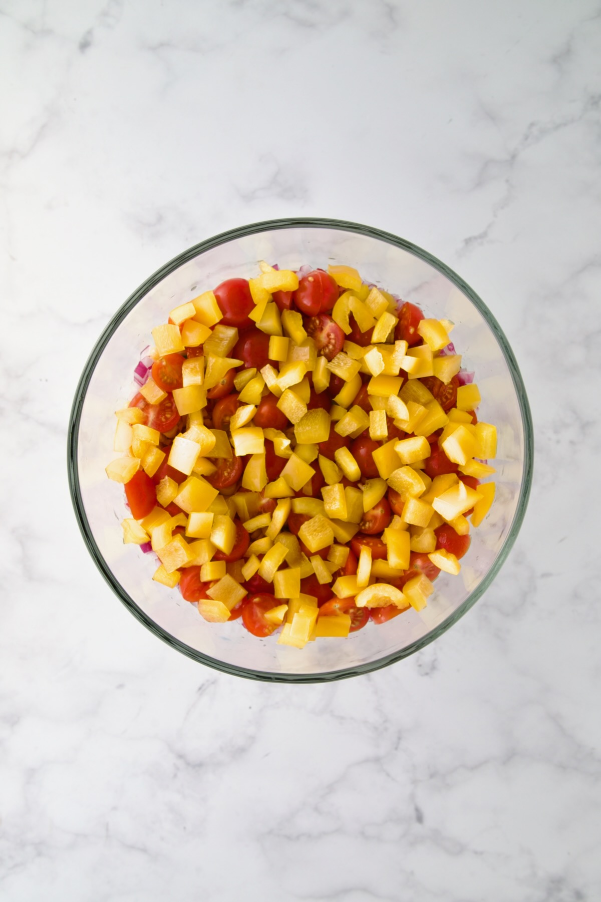 Diced yellow bell pepper over tomatoes in footed glass serving bowl.