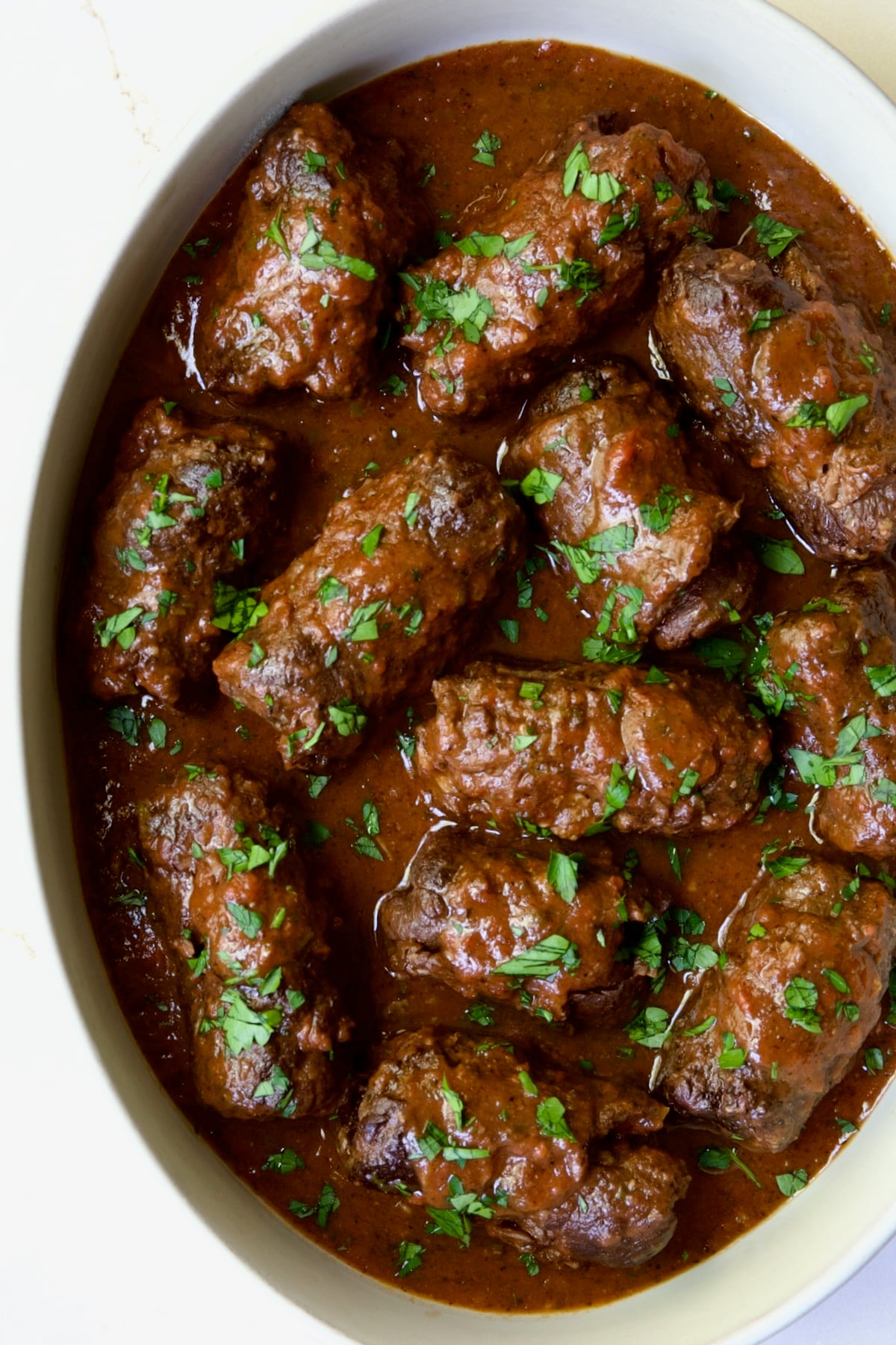 Fully cooked Beef Braciole in white oval dish garnished with fresh parsley.