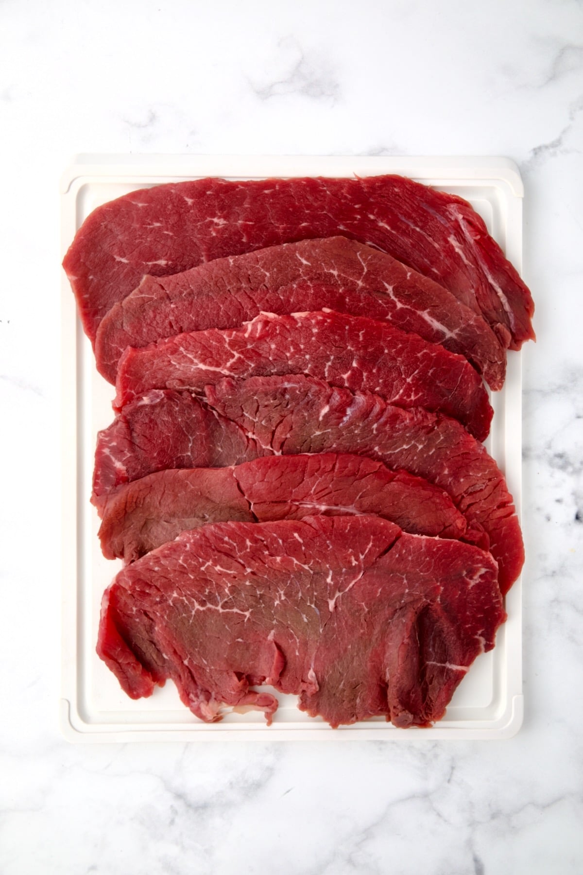 Six slices of beef top round on white plastic cutting board.