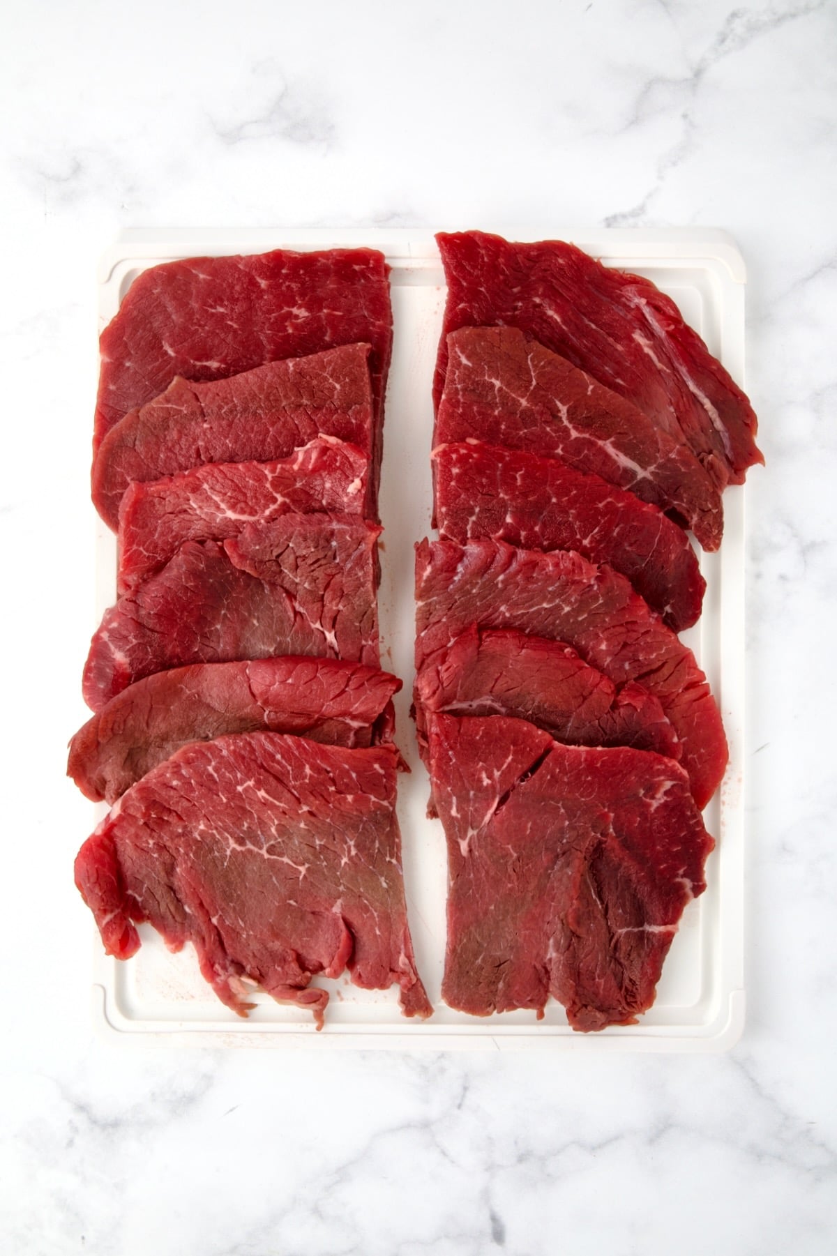 Six slices of beef top round on white plastic cutting board cut in half widthwise to create 12 pieces.