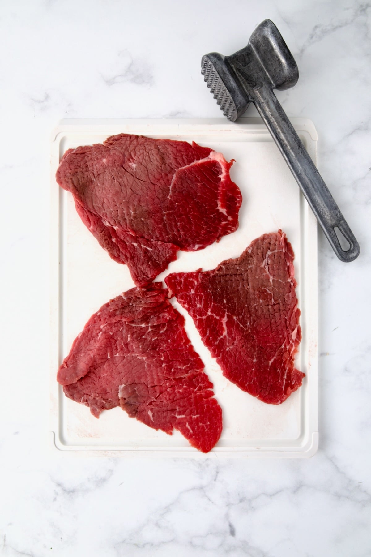 Three slices of beef top round on white cutting board with meat mallet to show how it's pounded.