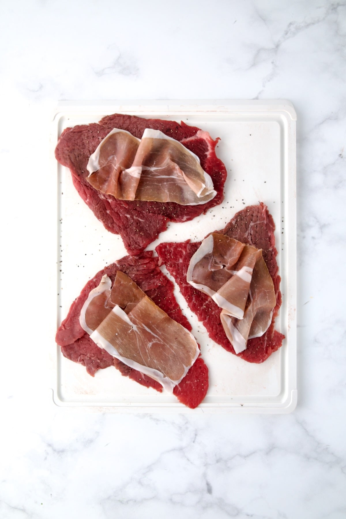 Three slices of beef top round on white cutting board seasoned with salt and black pepper with slices of prosciutto placed on top of the slices.