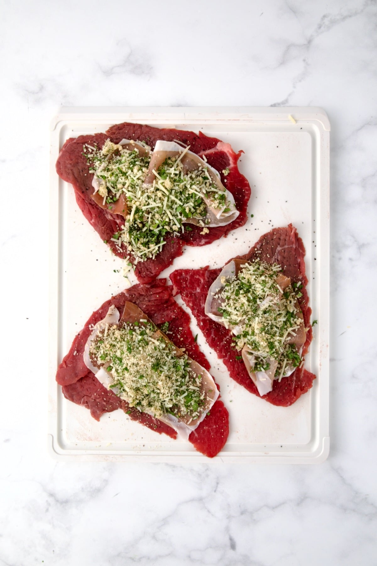 Three slices of beef top round on white cutting board with slices of prosciutto and the filling placed on top of the slices.