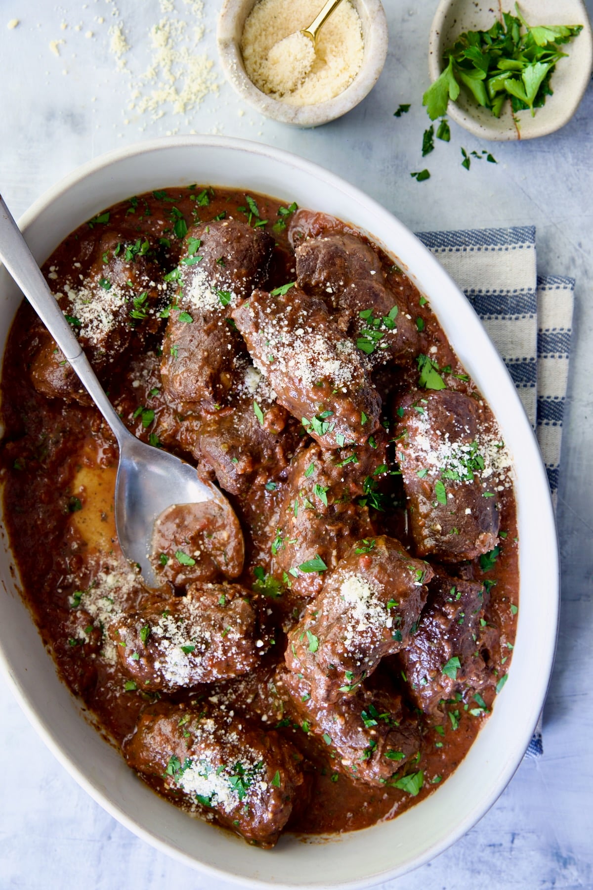 Beef Braciole in oval white serving dish with serving spoon on blue striped towel.