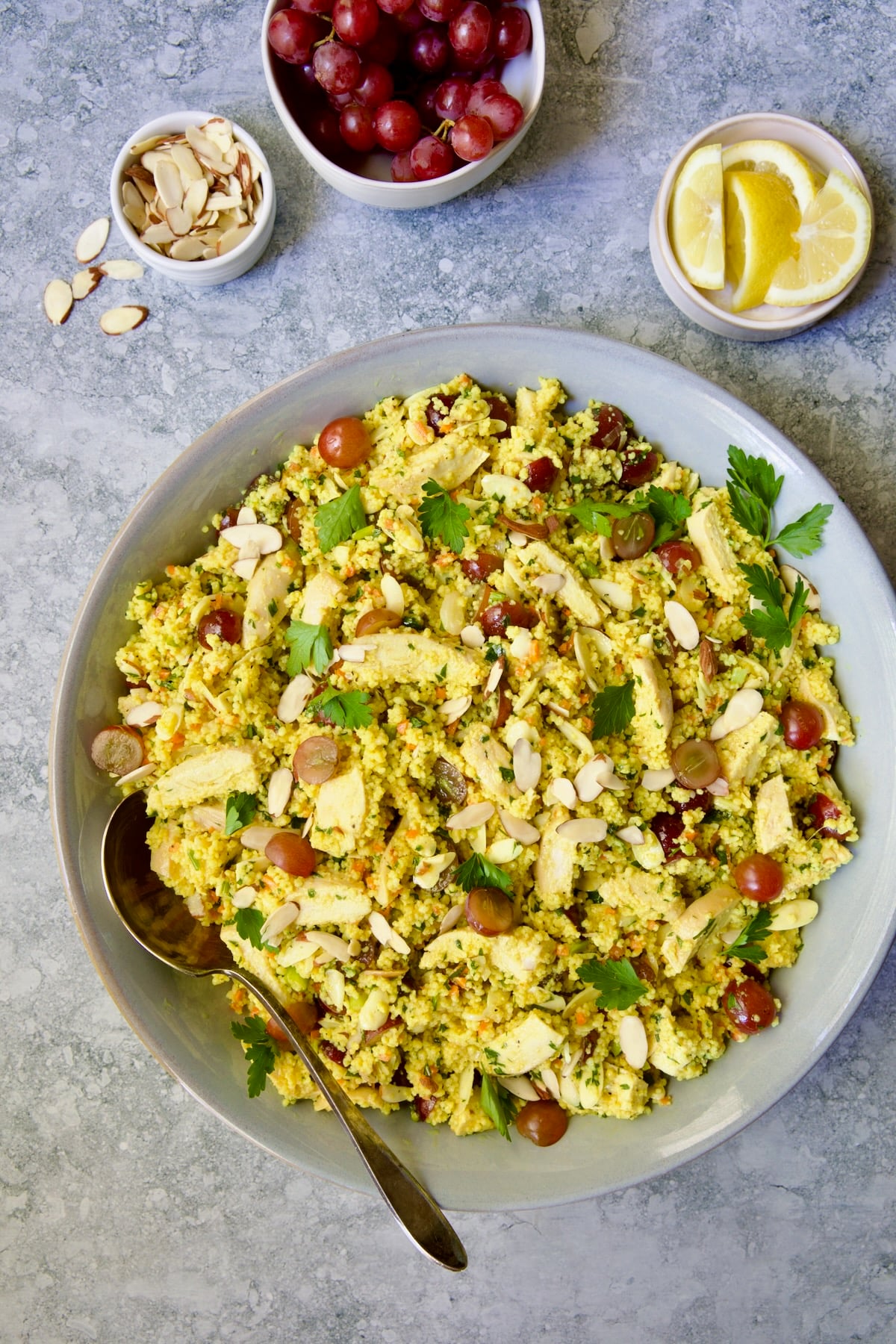 Chicken Couscous Salad in gray bowl with serving spoon with bowl of sliced almonds, whole grapes and lemon wedges.