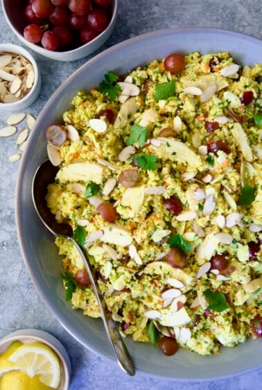 Chicken Couscous Salad in gray bowl with serving spoon with bowl of sliced almonds, whole grapes and lemon wedges.