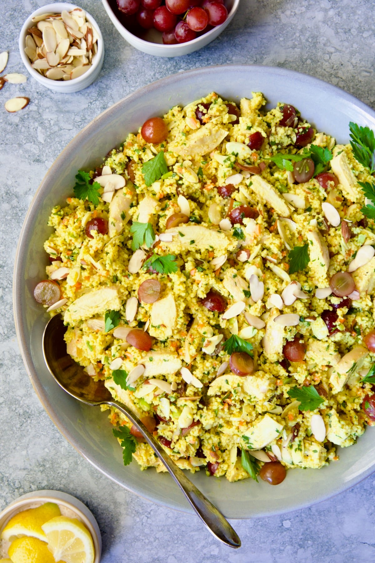 Chicken Couscous Salad in gray bowl with serving spoon with bowl of sliced almonds, whole grapes and lemon wedges.