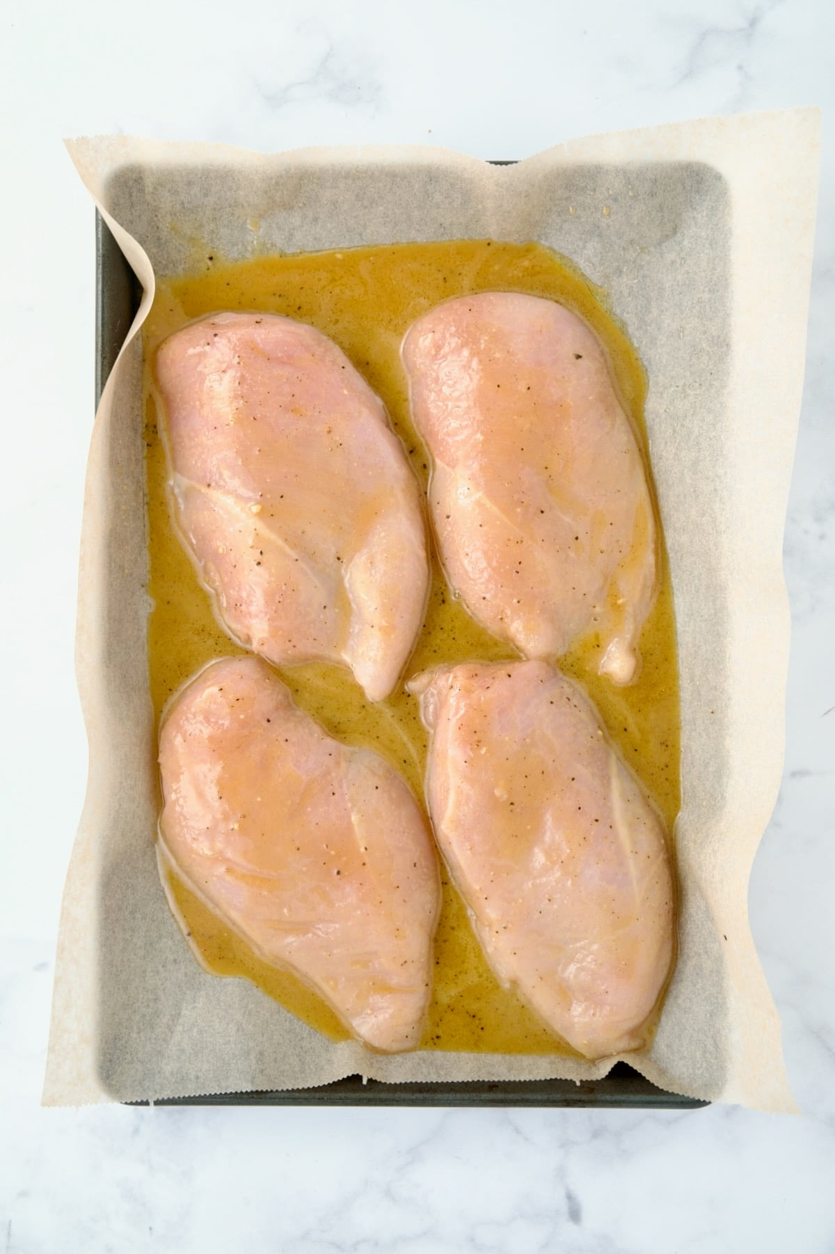 Four uncooked chicken breast halves on parchment paper lined rimmed baking sheet with honey mustard combination poured over them.