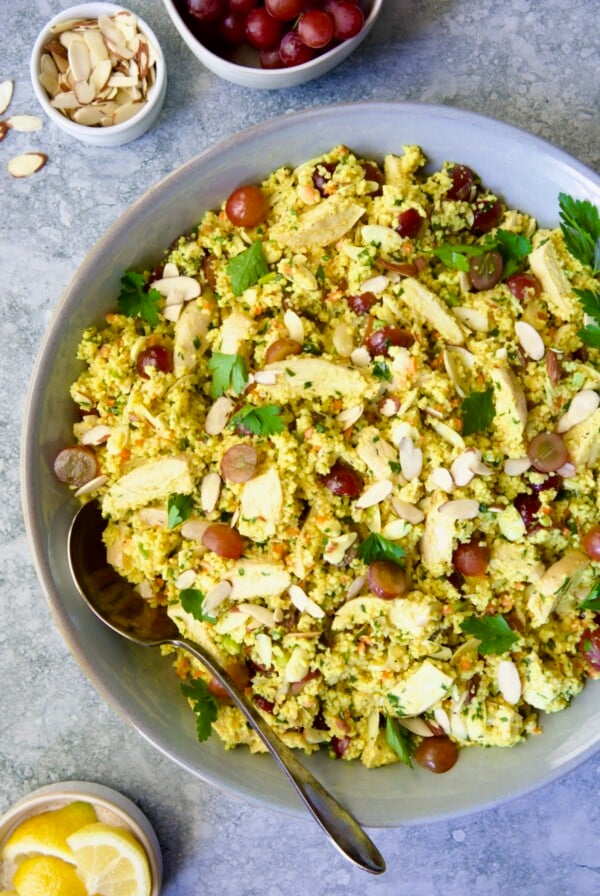 Chicken Couscous Salad in gray bowl with serving spoon with bowl of sliced almonds, whole grapes and lemon wedges.